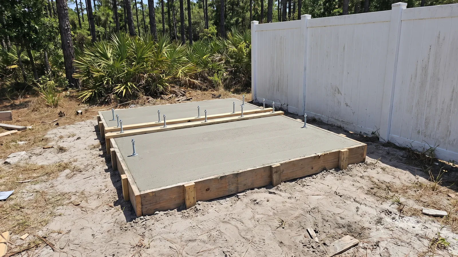 Reinforced concrete shed slab with anchor bolts set in a side yard beside a vinyl fence and pines